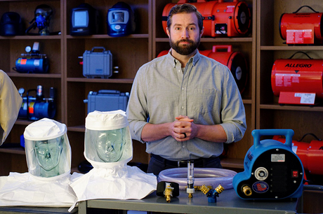 A man stands behind a table displaying protective hoods, hoses, and equipment. Shelves behind him hold red and blue industrial devices. He faces forward with hands clasped, wearing a light gray shirt.
