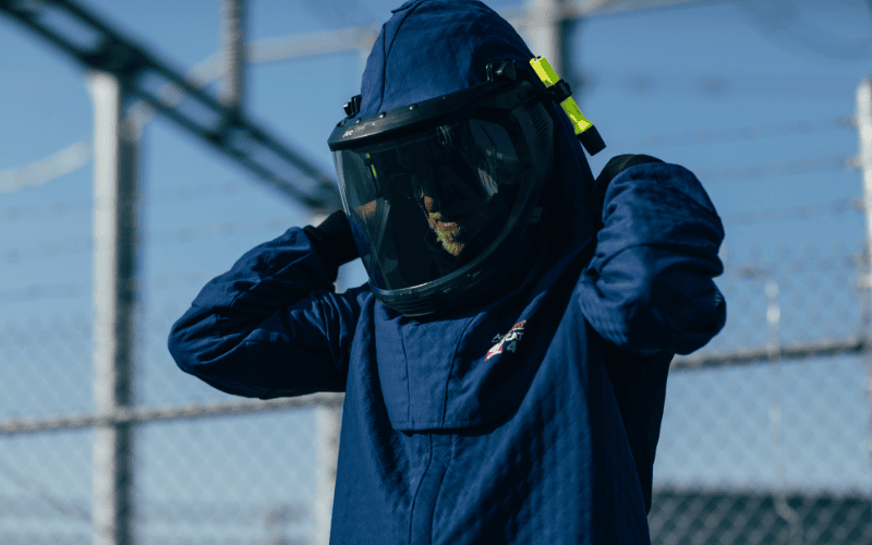 A person wearing a blue protective suit and helmet with a clear visor stands outdoors near a chain-link fence topped with barbed wire.