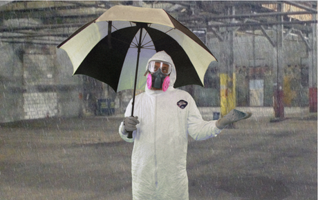 Worker wearing respirator and hazmat suit while standing in industrial facility holding an umbrella.