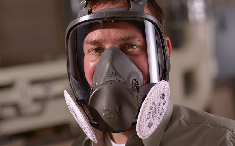 A man wearing a protective gray respirator mask with dual white filters smiles at the camera. The background is a blurred indoor industrial setting.