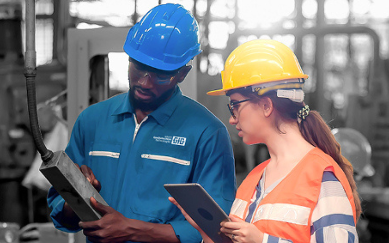 Two factory workers wearing helmets, one blue and one yellow, discuss a project while examining a device and a tablet, conveying collaboration and focus.