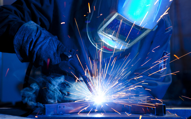 A welder in protective gear works with bright blue sparks flying. The intense light highlights the focused craftsmanship in an industrial setting.