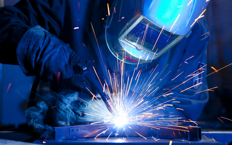 A welder in protective gear works with bright blue sparks flying. The intense light highlights the focused craftsmanship in an industrial setting.