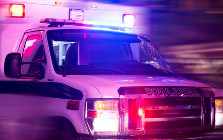 A speeding ambulance with flashing red and blue lights moves through a city street at night, suggesting urgency and action.