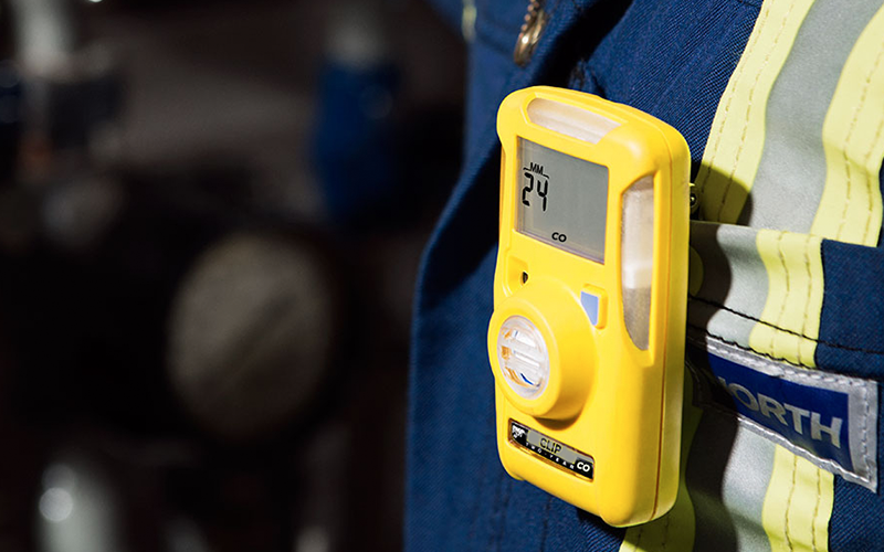 A yellow carbon monoxide detector clipped to a worker's blue industrial uniform displays "24" on its screen, indicating the detected CO level in the environment.