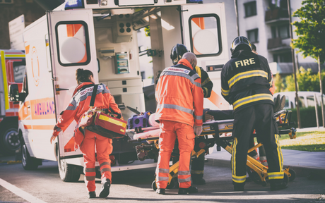Emergency responders in orange and black uniforms load a stretcher into an ambulance with urgency, conveying a sense of teamwork and focus.