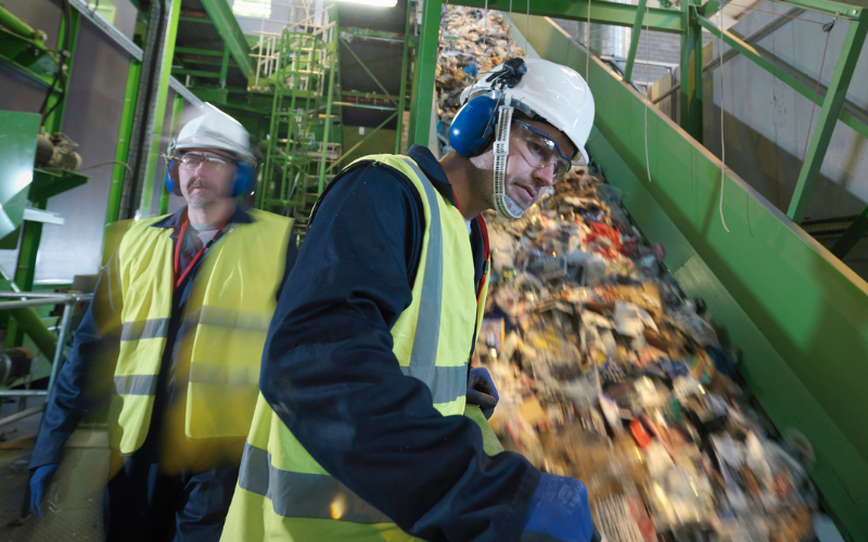 Two workers in safety gear, including helmets and ear protection, monitor a conveyor belt filled with mixed recyclables in a busy recycling facility.