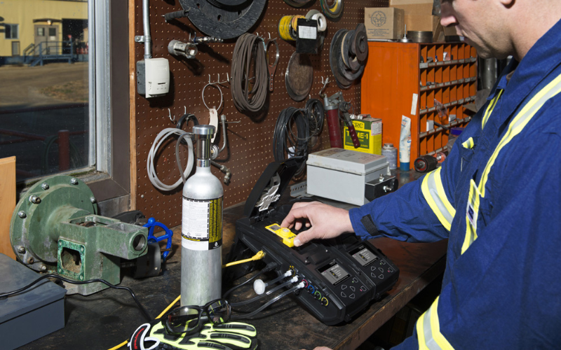 A person in a blue uniform operates gas detection equipment on a workshop bench with tools hanging on a pegboard. The workspace appears organized and functional.