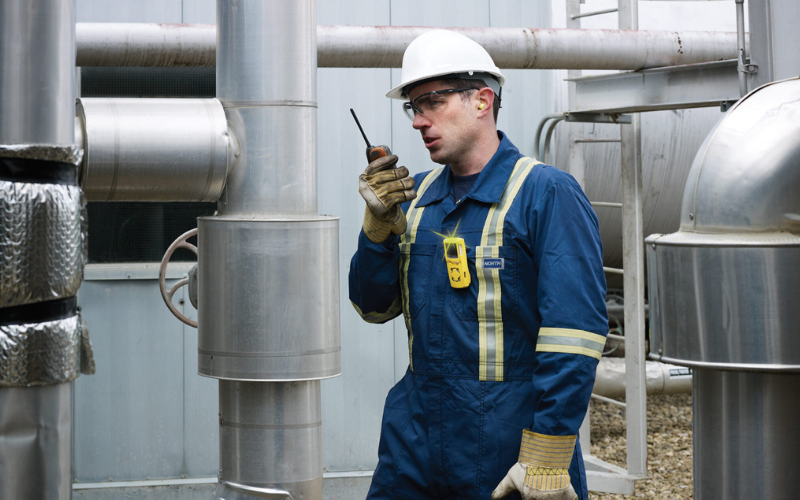 Worker in protective gear and helmet communicates via walkie-talkie amidst industrial pipes, conveying focus and diligence in a factory setting.