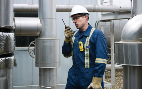 Worker in protective gear and helmet communicates via walkie-talkie amidst industrial pipes, conveying focus and diligence in a factory setting.