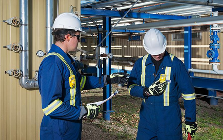 Two workers in blue safety uniforms and white helmets inspect industrial pipes outdoors. Each worker has a BW Clip gas monitor clipped to their chest.