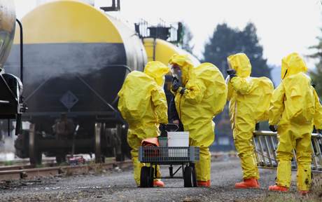 Four people in yellow hazmat suits stand near a rail track, inspecting large tanker cars. They appear focused and serious, suggesting an emergency response.