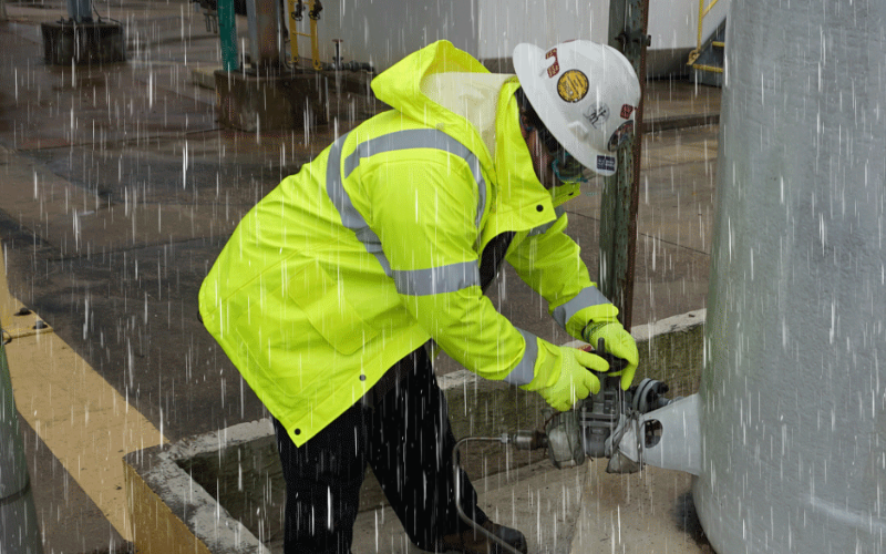 A worker in a bright yellow raincoat and hard hat checks equipment in heavy rain, conveying a focus on safety and diligence in challenging conditions.