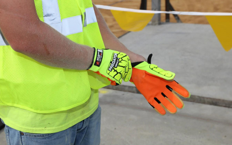 A person wearing a neon yellow safety vest adjusts a bright orange and yellow impact resistant glove. The background shows a construction area with caution tape.