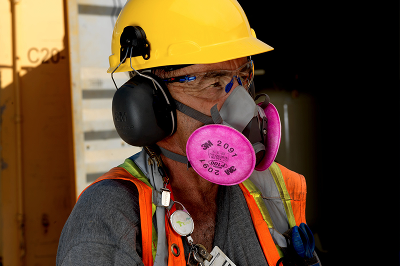 A worker wearing a yellow hard hat, safety glasses, a gray shirt, orange safety vest, and a respirator mask with pink filters stands in an industrial setting.