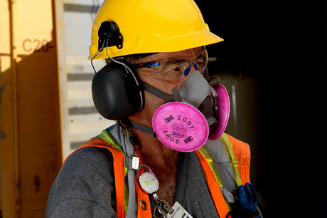 A worker wearing a yellow hard hat, safety glasses, a gray shirt, orange safety vest, and a respirator mask with pink filters stands in an industrial setting.