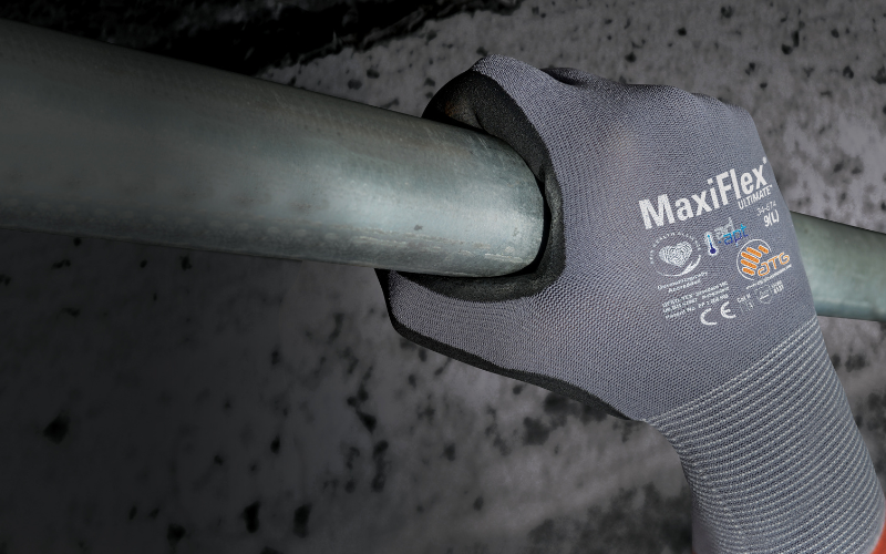 A gloved hand firmly grips a metal bar. The glove is gray with "MaxiFlex" branding, suggesting durability and grip in industrial settings.