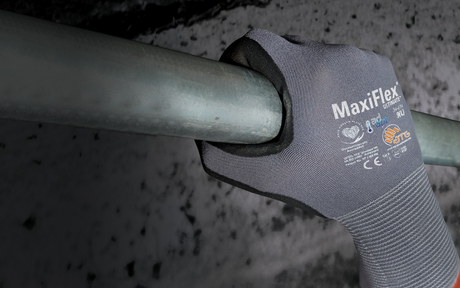 A gloved hand firmly grips a metal bar. The glove is gray with "MaxiFlex" branding, suggesting durability and grip in industrial settings.