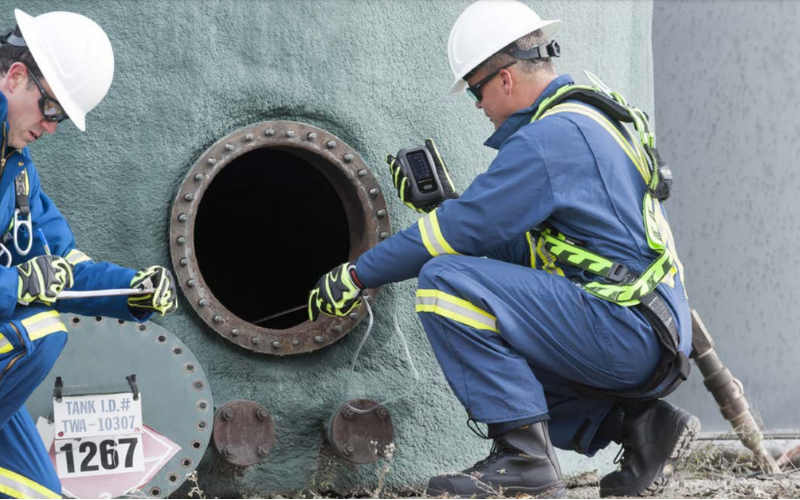 Two workers in protective gear examine an open industrial tank. Both wear blue safety suits, helmets, and gloves, conveying focus and caution.
