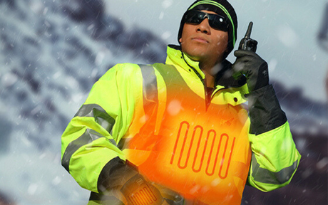 A person in a neon yellow heated jacket and sunglasses uses a walkie-talkie amid a snowy landscape. The scene conveys preparedness and resilience.