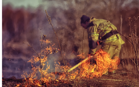 A firefighter battles a brush fire, surrounded by flames and smoke. The person wears protective gear, evoking urgency and bravery in the scene.