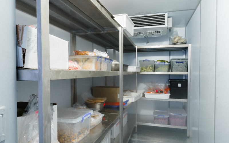 Industrial walk-in freezer with metal shelves holding labeled plastic containers of various foods. Cool, organized, and utilitarian atmosphere.