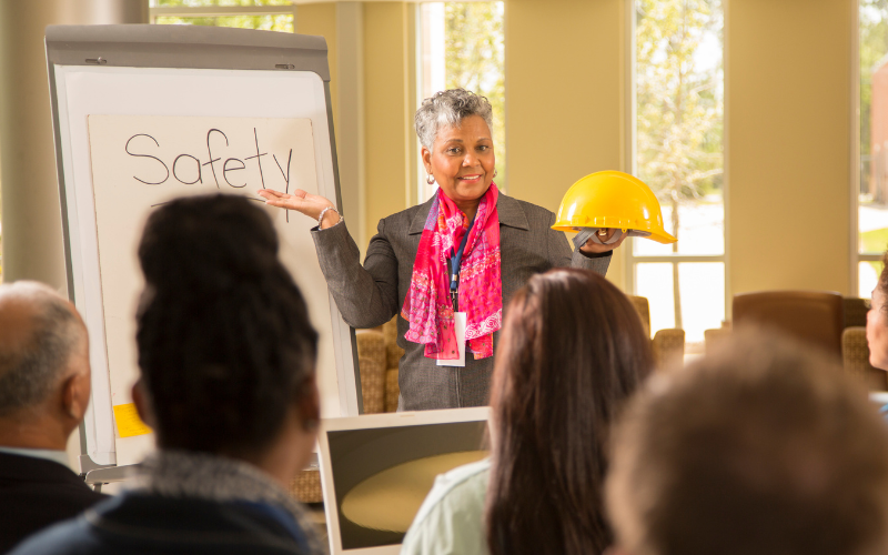 Woman giving safety presentation at office. Group of professionals sits and watches as she holds a construction hard hat with the word safety on a white board.