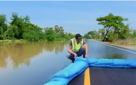 A person in a yellow vest secures blue sandbags on a flooded road, with lush greenery and a bright blue sky in the background, conveying urgency.