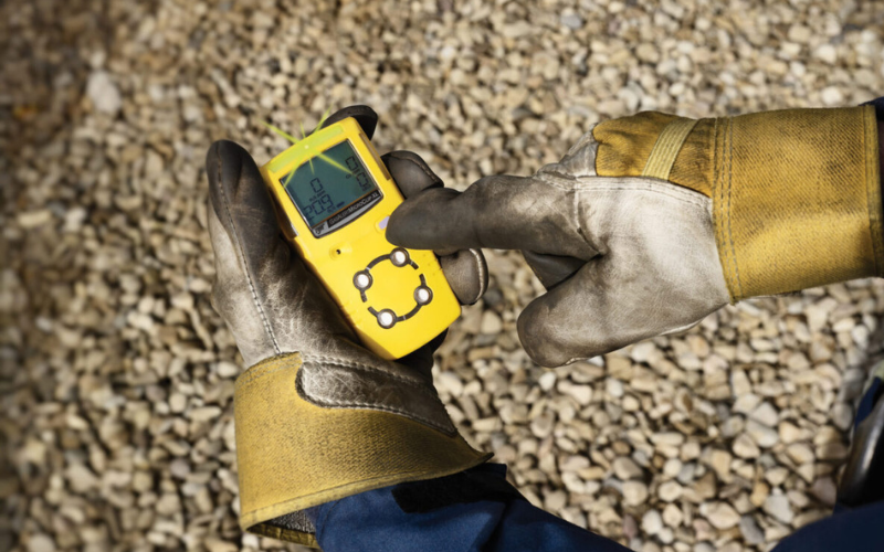 A person wearing rugged gloves holds a yellow handheld gas monitor with a screen displaying data. They stand over a gravel surface, suggesting an outdoor setting.