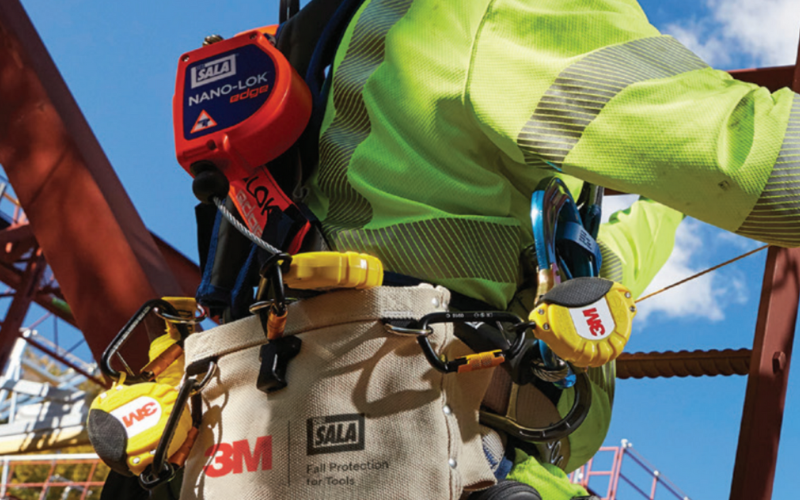A worker wearing a safety harness and bright green gear stands on a construction site, emphasizing safety with clear blue skies in the background.