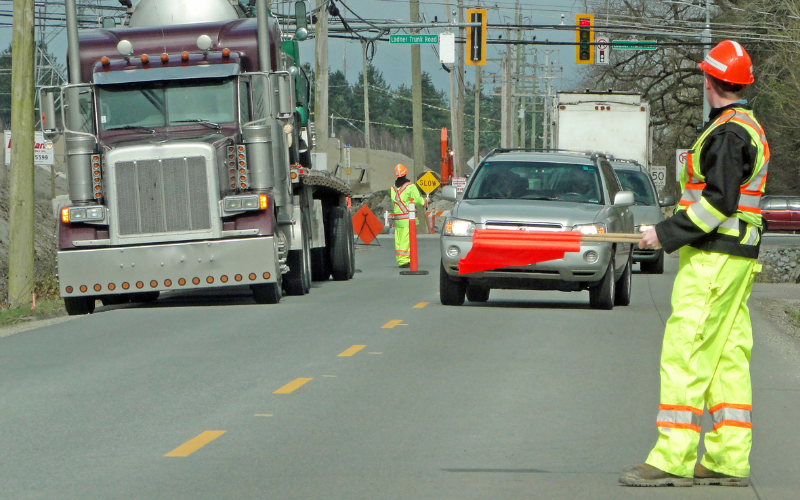 A construction worker in a neon safety suit and hard hat directs traffic with a red flag on a road. A large truck and a silver car are approaching.