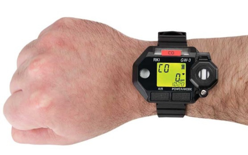 A rugged gas monitor wristwatch with a black strap on a wrist, displaying a bright yellow digital screen showing various numbers and icons.