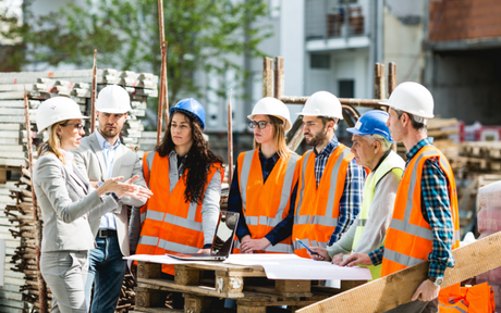 A group of construction workers in safety vests and helmets gather around a table discussing blueprints on a building site, conveying teamwork and planning.