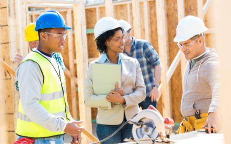 A diverse group of construction workers and a manager, wearing safety gear, discuss plans at a building site, conveying teamwork and collaboration.