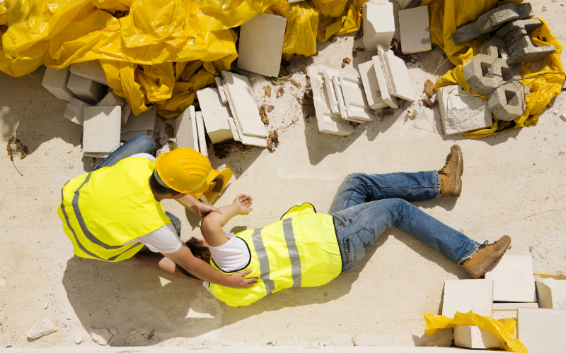 Construction worker has an accident while working on site. Another worker has hand next of them while they lay on the ground.