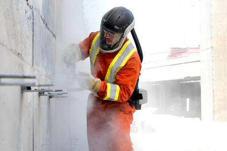 Worker grinding wall wearing protective gear including respirator, gloves, and coveralls.