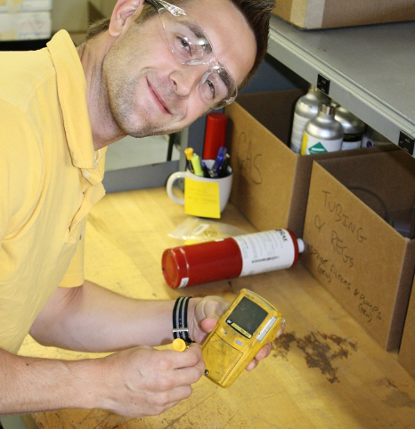 Worker wearing safety glasses stands over desk holding gas detector and smiles.