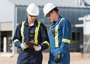 Two oil and gas workers in coveralls and hard hats with gas monitors clipped to their chests. One is holding a clipboard.