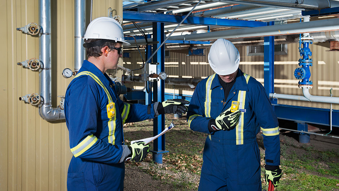 Two workers at an oil and gas site with pipes behind them. One is looking at a gas monitor clipped on his chest. Both wear PPE including helmets and coveralls.