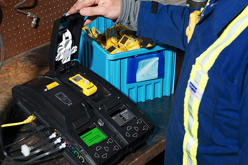 A person in a blue safety jacket operates a docking station for yellow gas detectors on a workbench, with a blue storage bin holding more detectors in the background.