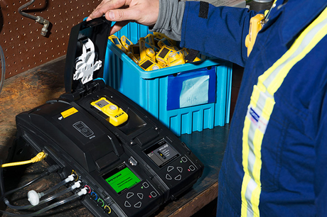 A person in a blue safety jacket operates a docking station for yellow gas detectors on a workbench, with a blue storage bin holding more detectors in the background.