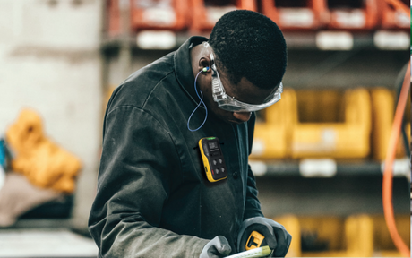 A worker in protective gear and goggles operates equipment in an industrial setting, surrounded by shelves with yellow containers, conveying focus and safety.