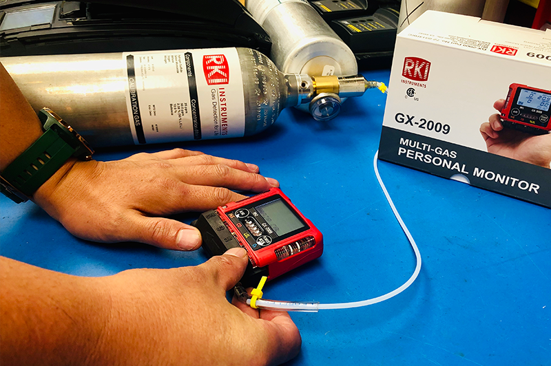 A person calibrates a red RKI GX-2009 multi-gas personal monitor using a gas cylinder and tubing on a blue surface. The product box and equipment are visible in the background.