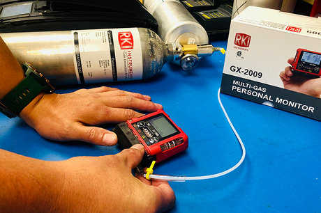 A person calibrates a red RKI GX-2009 multi-gas personal monitor using a gas cylinder and tubing on a blue surface. The product box and equipment are visible in the background.
