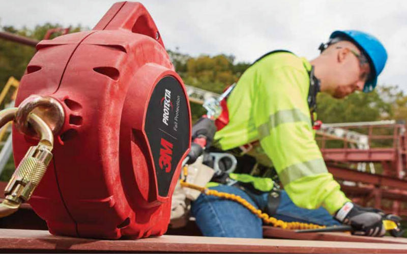 A construction worker in a blue helmet and green safety gear is using a harness on scaffolding. A red 3M fall protection device is in the foreground.