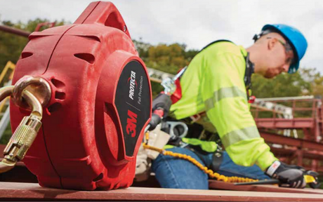 A construction worker in a blue helmet and green safety gear is using a harness on scaffolding. A red 3M fall protection device is in the foreground.
