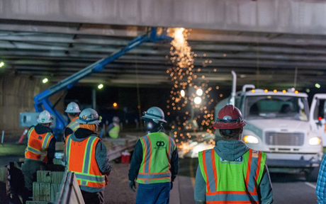 Construction workers in safety gear and vests stand under a bridge at night. Sparks fly from welding, while a truck is parked nearby, creating an industrious scene.