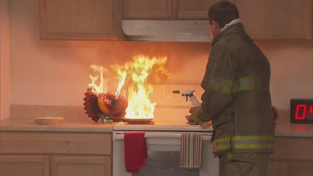 Person dressed in firefighter jacket putting out a fire on a kitchen stove.