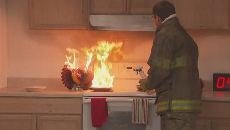 Person dressed in firefighter jacket putting out a fire on a kitchen stove.