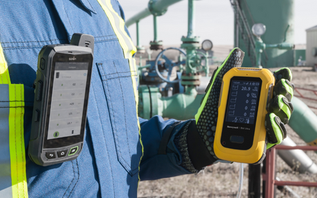 A worker in blue protective gear holds a yellow handheld device displaying data. Another device is clipped to their chest. Industrial pipes are in the background.